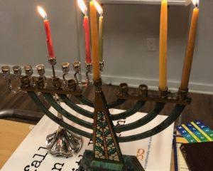 Two chanukiyot (menorahs) stand on some SVARA materials, lit for the second night of chanukah (one center candles, two candles on the right).