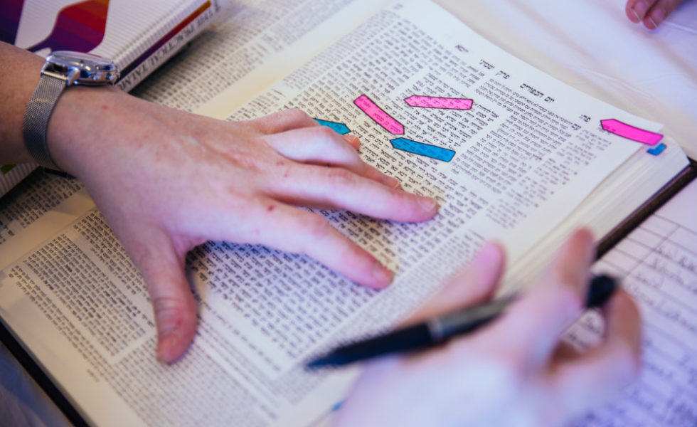 A left hand points at a line in the Talmud, while a right holds a pen.