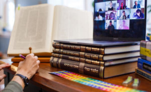 A computer sits on top of a stack of Talmuds, with a Zoom meeting open. Many faces are visible in the Zoom window. Next to the computer, a person studies from a Talmud, their hand holding a pencil.