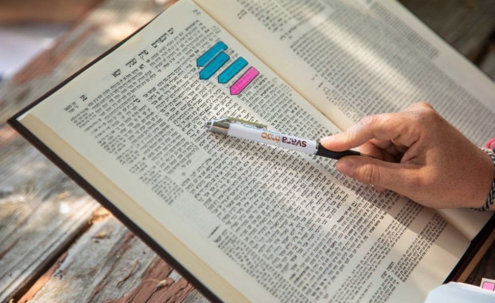 A person points a SVARA-branded pen at a line of Talmud.