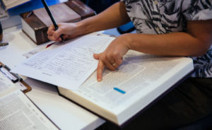 A person's left hand points at their Talmud, while the right takes notes on a piece of paper.