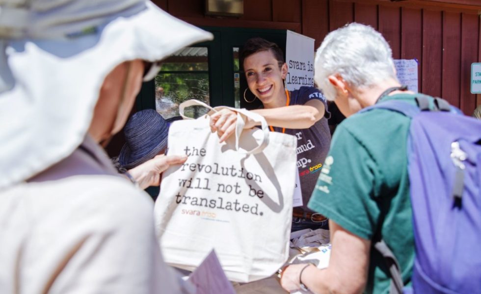 Rabbi Monica, smiling, hands a SVARA tote bag to someone outside the frame. The tote bag reads, "The Revolution Will Not Be Translated."