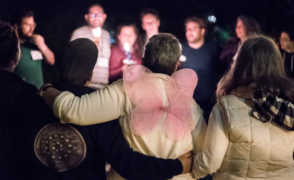 A group of SVARA-niks stand in a circle around a burning havdalah candle. In the foreground, a SVARA-nik is wearing fairy wings on their back.