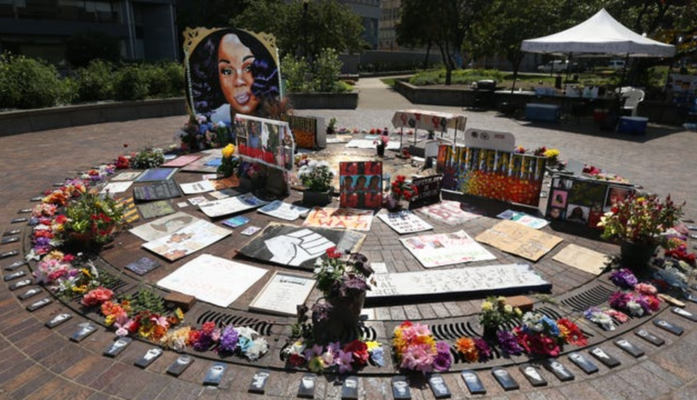 An outdoor memorial for Breonna Taylor, arranged in a circle. There are flowers, a large portrait of Breonna, and various smaller artpieces, including a Black Power fist.