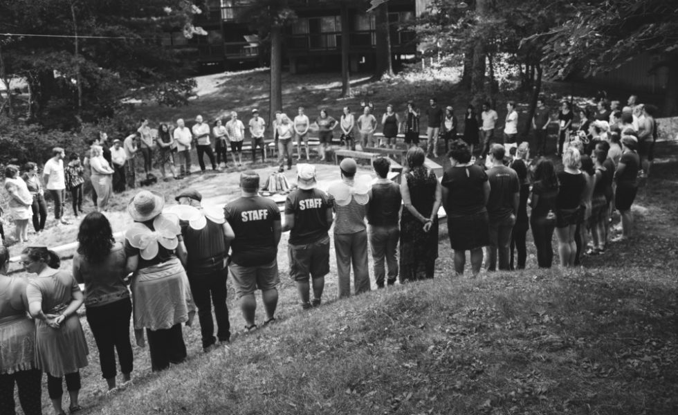 A black and white photo of a large circle of SVARA-niks, all standing outside, around a campfire that is not burning.
