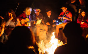 A group of SVARA-niks, a few of whom are playing guitars, sit around a campfire at night.