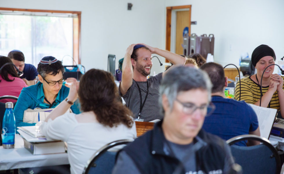 A large group of SVARA-niks are sitting at tables in a bet midrash. In the center, one SVARA-nik, wearing a grey shirt, has their hands on their head and a smile on their face.