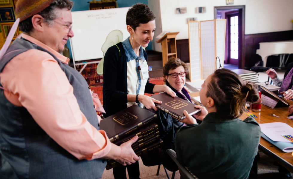 On the left, there are two standing SVARA-niks, and on the right, two seated. Rabbi Monica, standing center, is handing a Talmud to one of the seated learners.