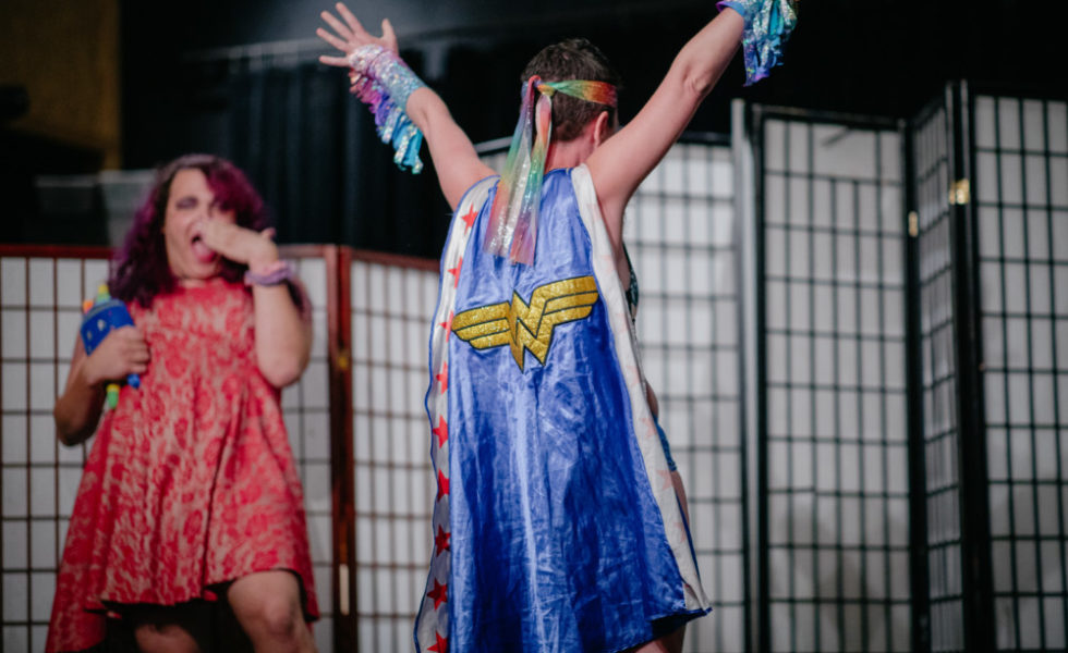 A SVARA-nik stands with their back to the camera, showing off a glittery blue cape with Wonder Woman's logo on the back. Their arms are up in the air, and they are wearing glittery streamers on their wrists. Another SVARA-nik, off to the left, looks on, amazed.