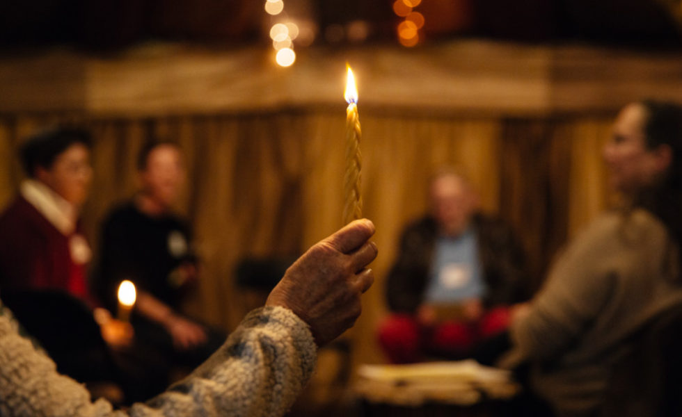 In the foreground, a hand holds a beeswax, golden havdala candle, which is lit. In the background, which is blurred, four SVARA-niks stand around the candle.