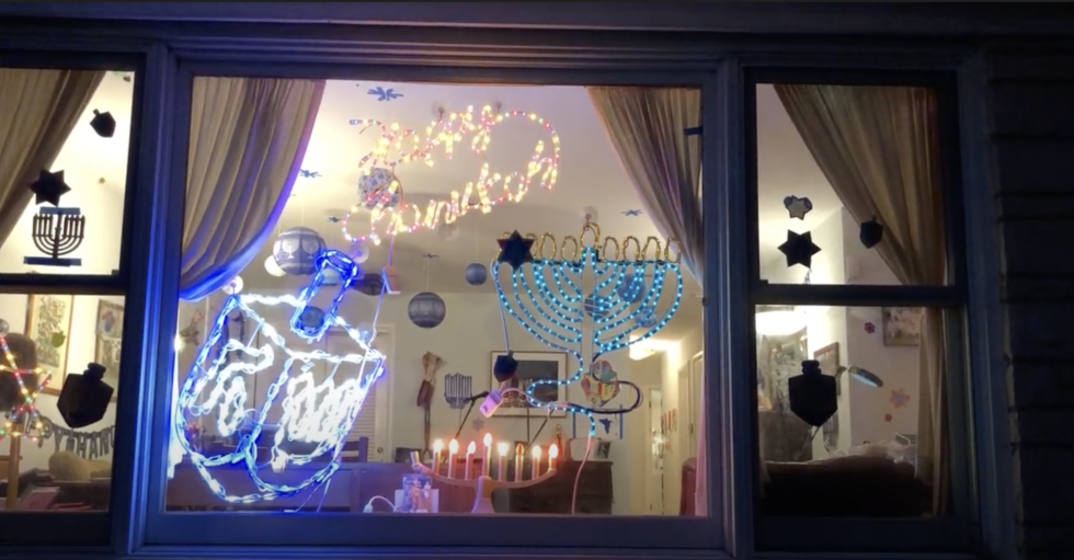 The window of someone's home, taken outside and at night. The window is decorated with several chanukah messages - a light up dreidel, a lit up message saying happy chanukah, an outline of a menorah, and at the bottom, on the windowsill, a menorah, its candles burning.