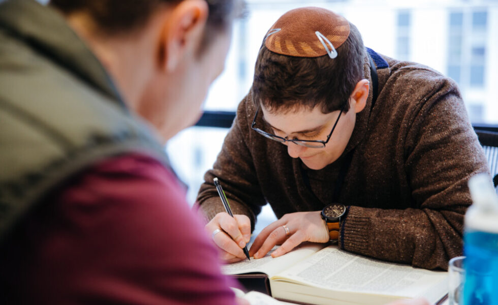 Rabbi Becky, wearing a brown sweater and a lighter brown kippah, leans over a Talmud, writing notes.