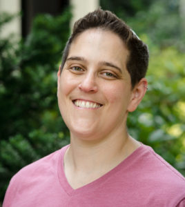 Rabbi Becky is visible from the shoulders up. He is wearing a pink v-neck t-shirt, and is smiling with his head tilted slightly to the right. He is standing in front of a blurred bushy background, with some flowers visible.