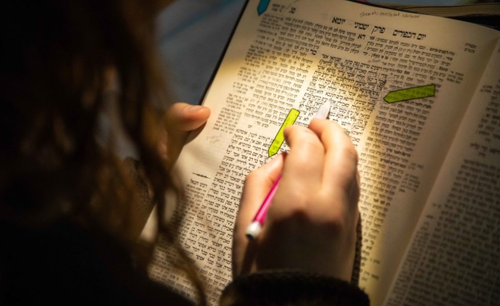 Someone's hand, holding a pen, points to a line in the Talmud. The Talmud is lit up by a lamp, so that part of the page is illuminated and part of it is in shadow. The person's long, wavy brown hair is visible on the left of the photo.