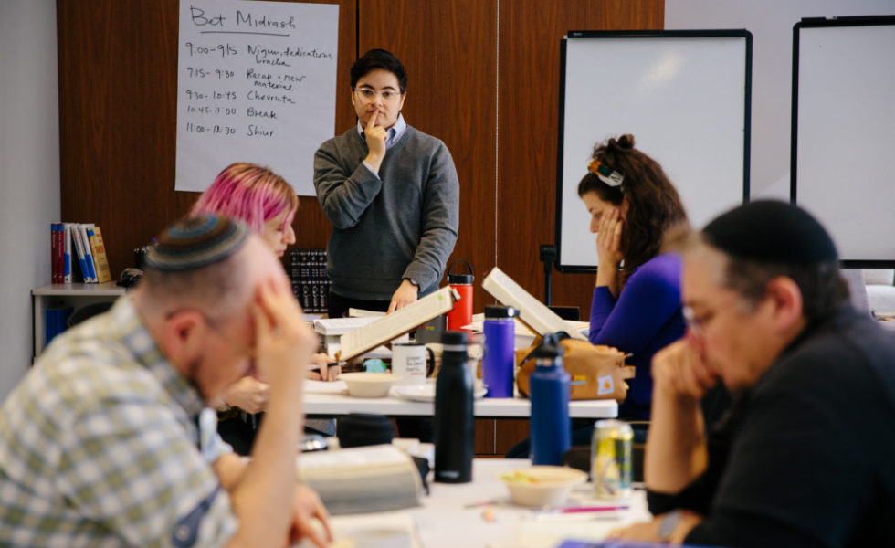 Laynie stands at the back of the bet midrash, their finger on their lips in a thinking pose. Bronwen and Jhos sit at tables in front of Laynie, learning with their respective chevrutas.