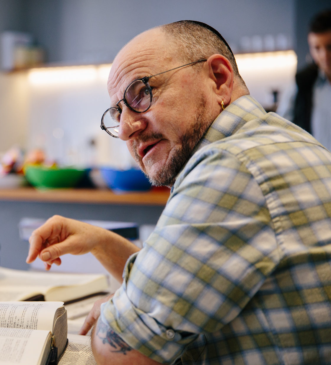 Jhos Singer sits in the bet midrash. There is a volume of Talmud in front of him and he glances towards someone standing outside of the frame. Jhos is wearing a checkered button-up shirt with the sleeves rolled to his elbows.