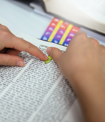A person places both of their hands on a page of Talmud. Above the page is a rainbow Alef-Bet learning resource.