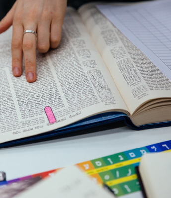A person places their hand on a page of Talmud. A rainbow alef-bet learning resource is placed below the page.