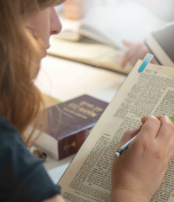 A person places their pencil on a page of Talmud. They are wearing a turquoise shirt, and a dictionary is placed next to them in the bet midrash.