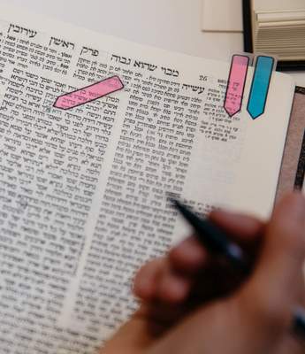 A page of Talmud is centered in the frame. There are three sticky notes placed on the page. A person's right hand can be seen in the foreground of the image.
