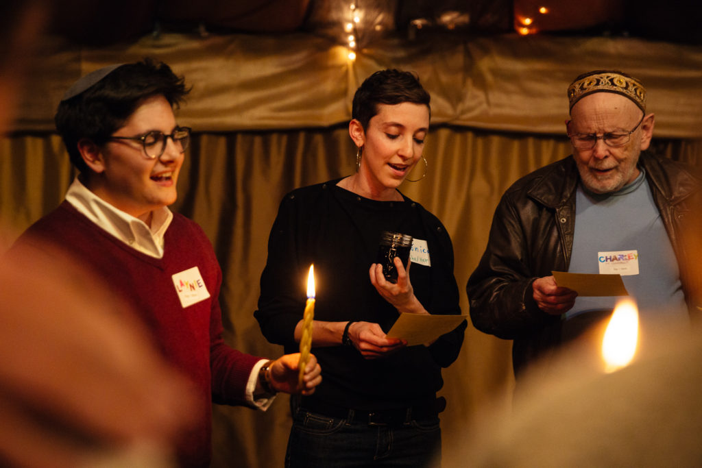 Three people stand in a circle to welcome in havdalah. They are smiling and singing together, and a havdalah candle burns towards the center of the image.