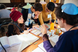Four people are gathered around a table in the bet midrash. Two of them are seated in chevruta and two are standing next to them. Everyone is focused on the text in front of them, and it appears that there is deliberation and deep attention being paid to the text.