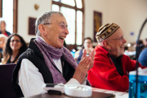 Two SVARA-niks sit next to one another in the bet midrash. They are both smiling, clapping, and looking ahead towards someone out of frame.