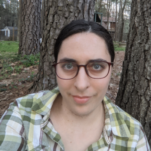 Esther is standing outside in front of trees. She is wearing a green plaid shirt and glasses with dark frames.