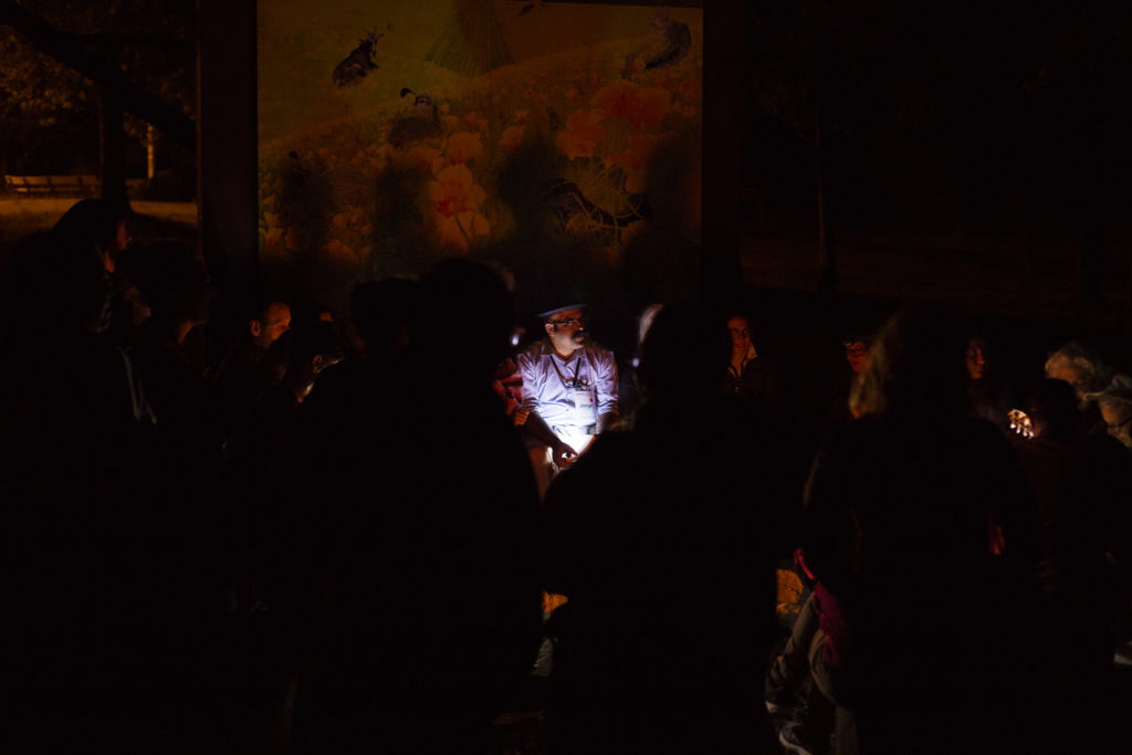 A group of SVARA-niks gather in the darkness around the dim light of a fire. One person's face is illuminated by a flashlight. They look to the right towards someone speaking. The shadows of the folks sitting are cast on the wall behind them.