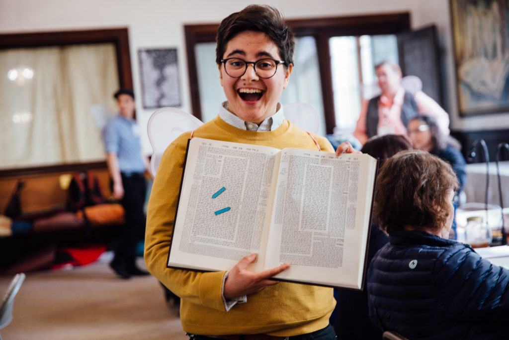 Laynie stands in the bet midrash holding a volume of Talmud. They are wearing a yellow sweater and dark eyeglasses.