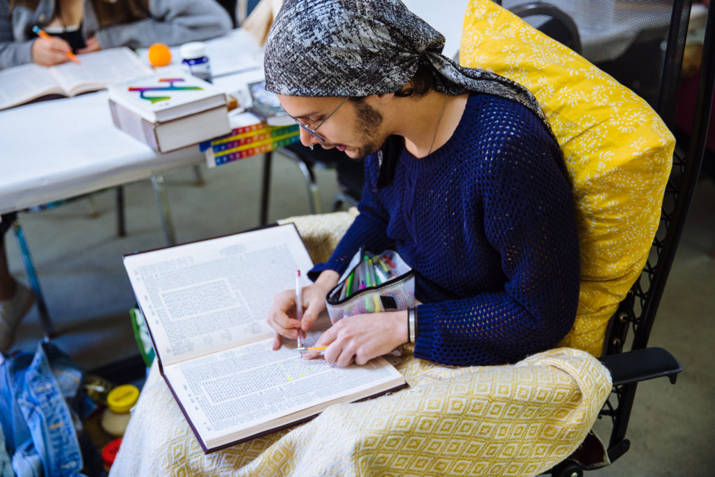 A SVARA-nik sits on a chair with a bright yellow pillow and a blanket over their legs. They are leaning over an open Talmud in their lap, which they are annotating with a pen and pointing to. They are reading intently from the text.