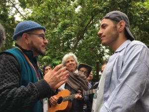 Two learners stand outside at Queer Talmud Camp. They are facing one another, and they are surrounded by other learners who appear to be gathered in a circle around them.