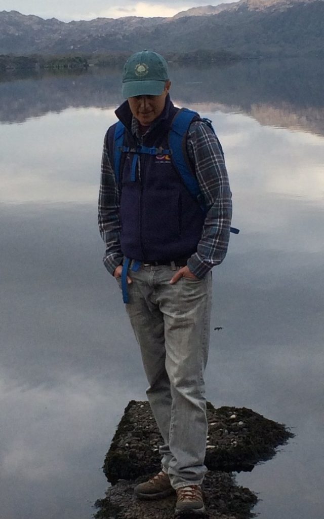 Leykn stands in front of a lake ringed by mountains, visible from the chest up. They are wearing a green baseball cap and a blue body warmer over a flannel.