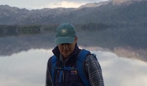 Leykn stands in front of a lake ringed by mountains, visible from the chest up. They are wearing a green baseball cap and a blue body warmer over a flannel.