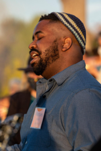 Marques is standing in the golden light of a setting sun, with their mouth open in song. They are wearing a blue collared shirt and an orange name tag.