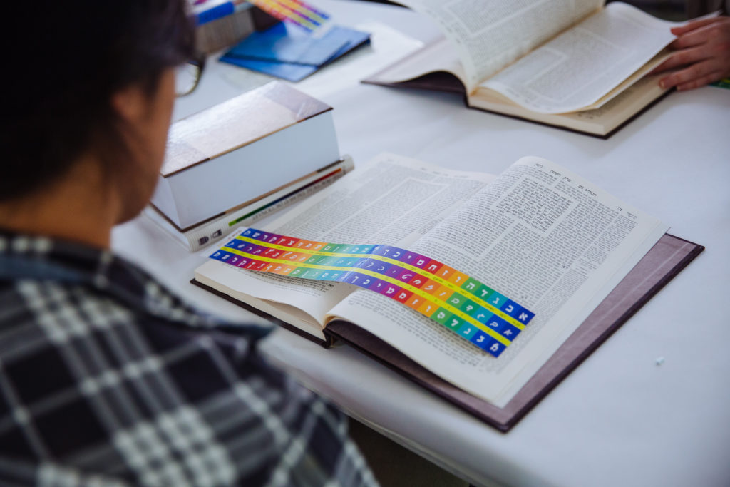 A volume of Talmud sits open on a table. A learner can be seen sitting next to it. There is a rainbow alef-bet tool placed across the pages.