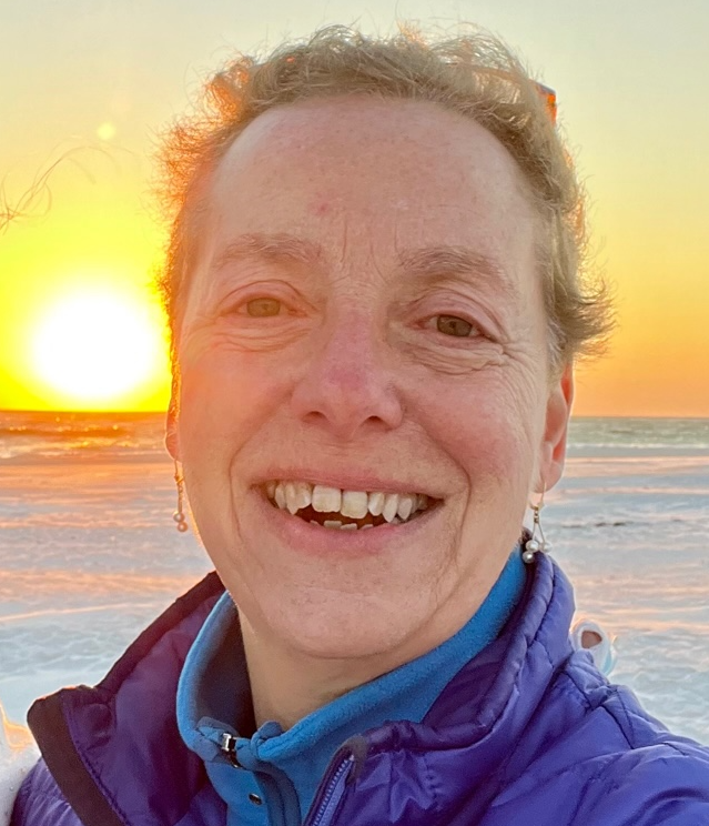Sandy, visible from the shoulders up, smiles in front of a sunset over the sea. She is wearing a purple jacket over a blue turtleneck.