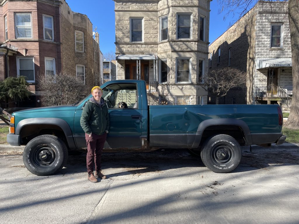 Maggie is standing in front of their green pickup truck. They are wearing a yellow hat and a green winter coat. Their dog can be seen sitting in front of the steering wheel. It is a bright blue winter's day in Chicago.