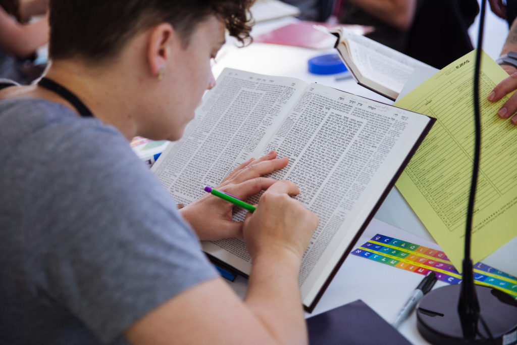 A learner is seated in the bet midrash, pointing to a section of a page of Talmud. They have a pencil in one hand, and the table beneath them has a lot of learning resources scattered about.