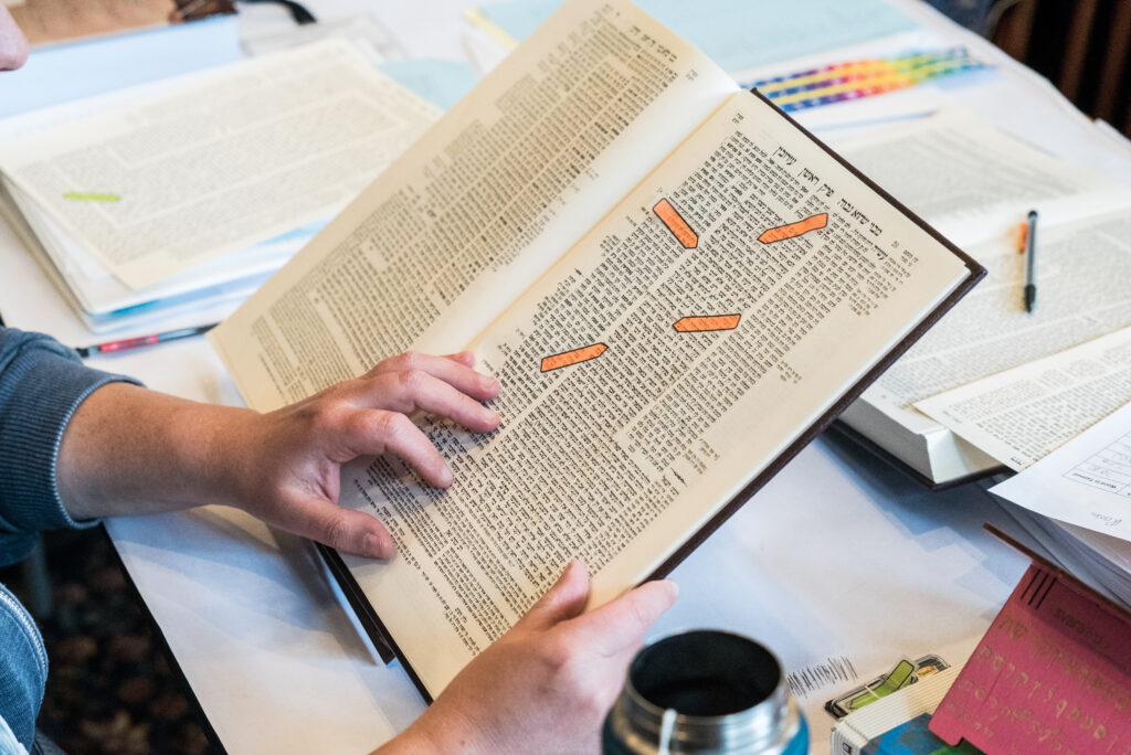 A learner reads from a masechet resting on a shtender. There are orange sticky-arrows scattered across the page, indicating where a certain line of Talmud starts and ends.