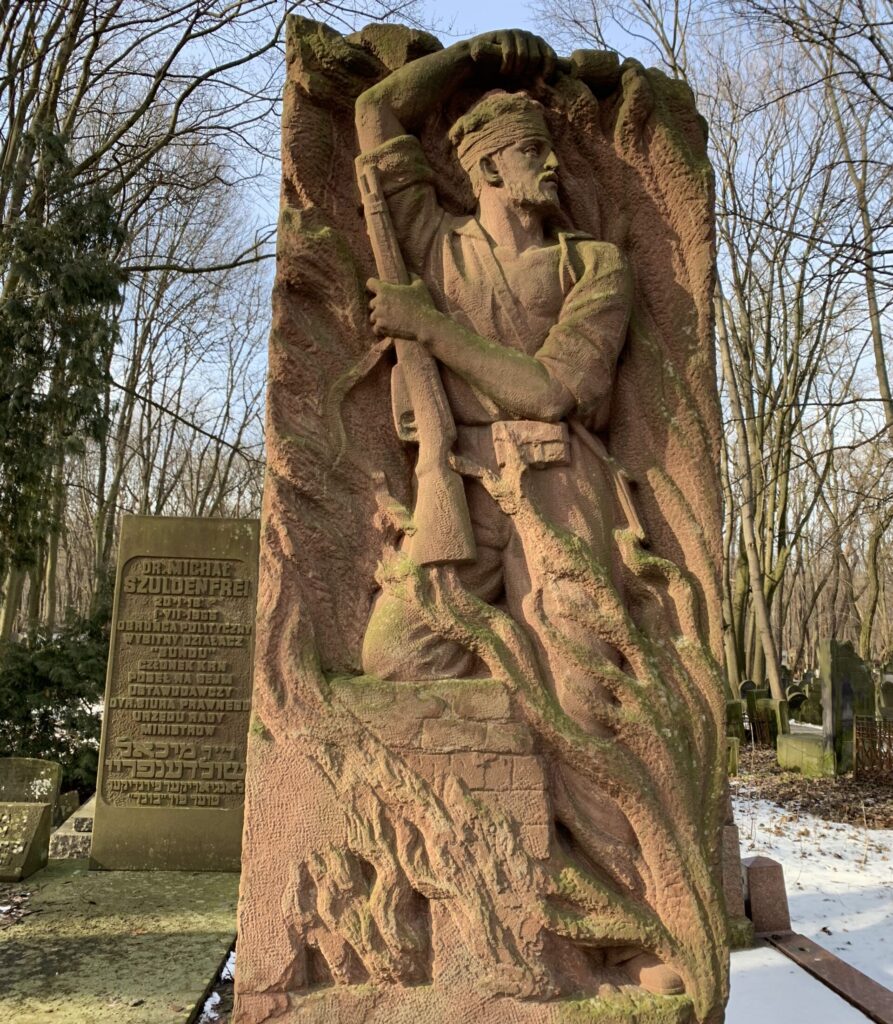 A stone, rectangular monument carved with a sculpture of a Bundist fighter marching amidst flames and holding a rifle. In the background is a gravestone with Polish and Hebrew writing, and a snow-covered path.