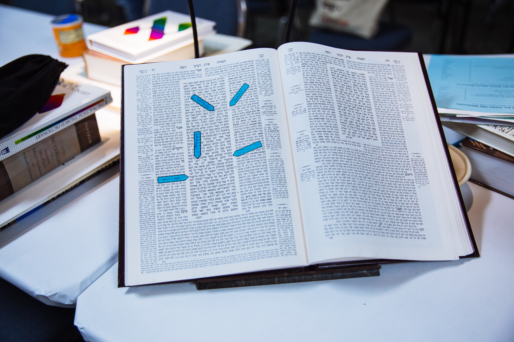 A Talmud sits on a table, propped up by a bookstand, open to a page. There are small, blue stickies marking the place on one of the pages.
