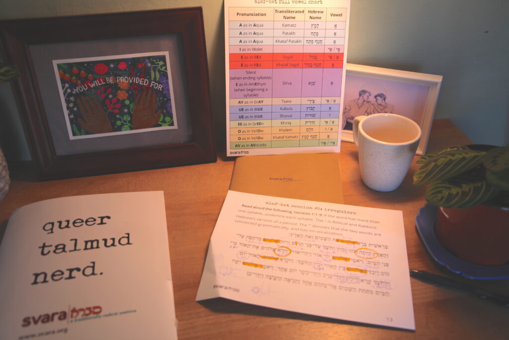 A close-up image of someone's Alef-Bet learning materials. There is a colorful alef-bet sheet that lists each letter and its definition, as well as a folder that says "queer talmud nerd", and a worksheet that has been underlined and highlighted.