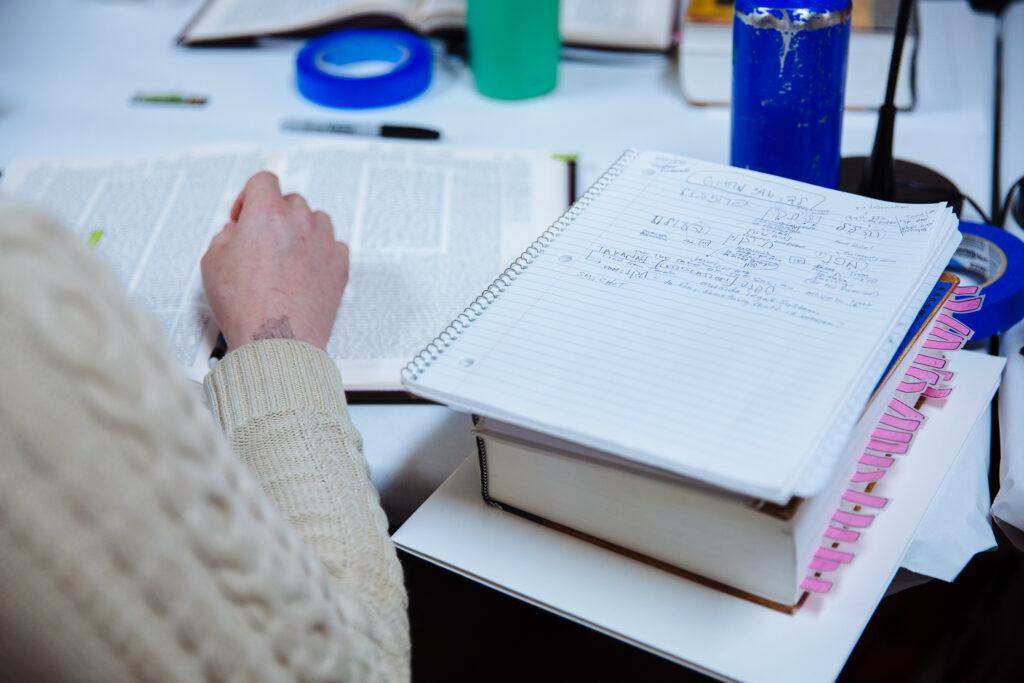 A learner sits in the bet midrash with a stack of materials beside them. Their notebook, as well as a masechet full of pink sticky notes, sits to the right of them.