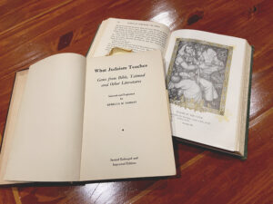 Two books are open on a dark wooden table. The first is opened to its title page, which reads, "What Judaism Teaches by Rebecca Namiat: Gems from Bible, Talmud, and other Literatures." The other book is not identifiable, but has an intricate wood-carved print on its page.