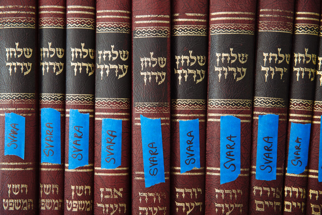 A collection of many volumes of Talmud. They are placed on a shelf, one after the other, and all of them have a blue piece of tape on the binding that reads, "SVARA".