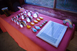 A table of learning materials is set up in the SVARA bet midrash. There are laminated dafs, or pages of Talmud, alongside magnifying glasses, highlighters, unicorns, and rainbows. The table cloth is magenta and light from a nearby window falls across the table.