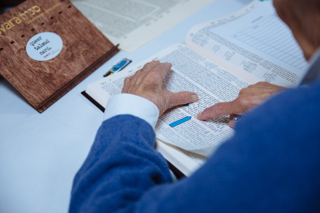 A SVARA-nik is reading a page of Talmud in the bet midrash. The image is zoomed in on the learner's hands which rest on the page in front of them. They are wearing a soft blue sweater, and a SVARA branded shtender sits in front of them.