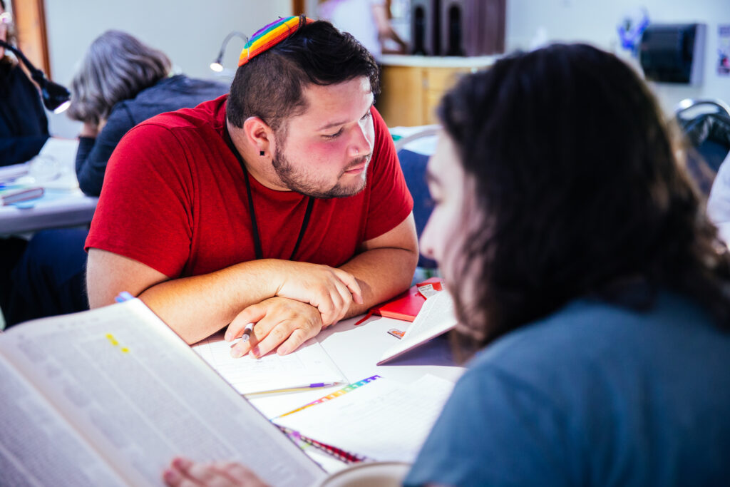 A SVARA-nik is peering over the table at the Talmud learning materials in front of them. They are wearing a red t-shirt and a rainbow kippah.