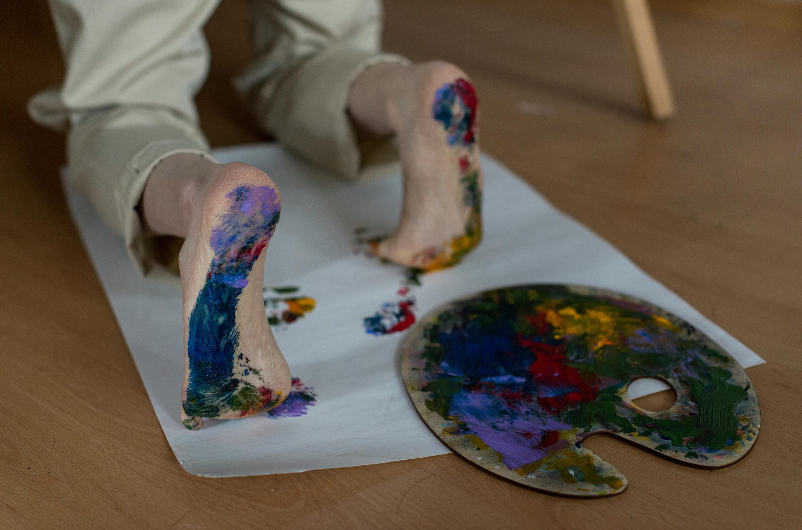 Someone makes art using paint on their feet. They are kneeling and stepping on a large sheet of white paper.